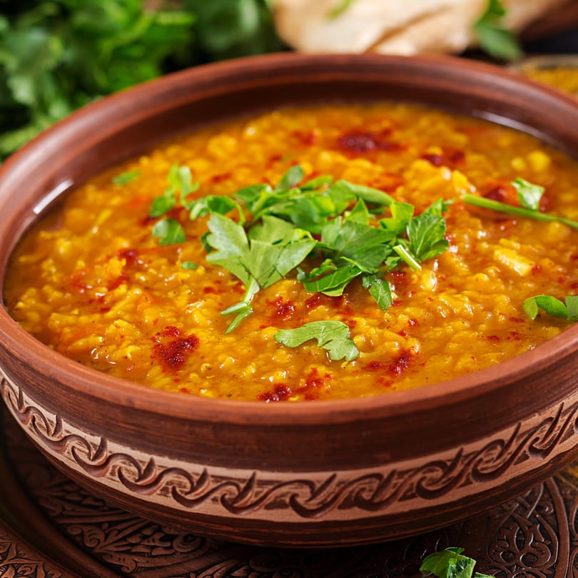 A close-up of a brown bowl filled with yellow Indian Nepali dal, garnished with red spices and fresh green herbs.