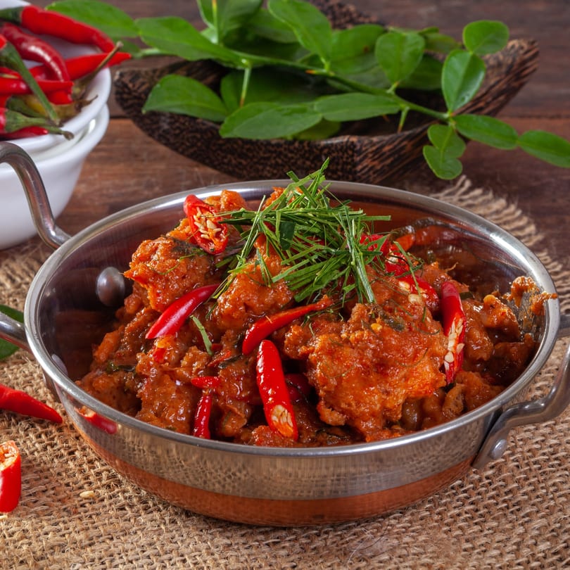 Spicy Indian curry with red chilies and green herbs in a copper-bottomed bowl on burlap.